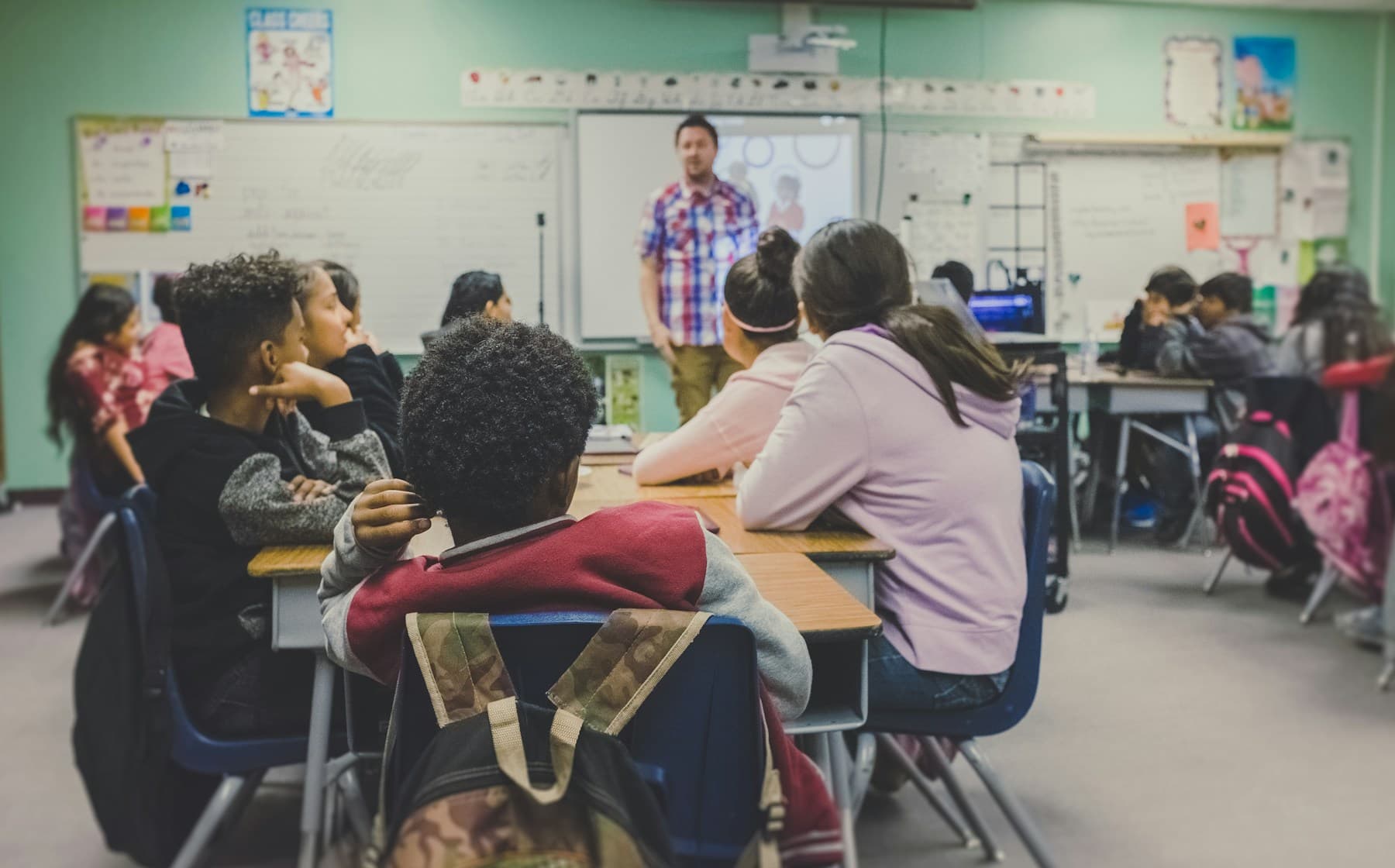 A classroom in session — students at desks, teacher presenting at the whiteboard.