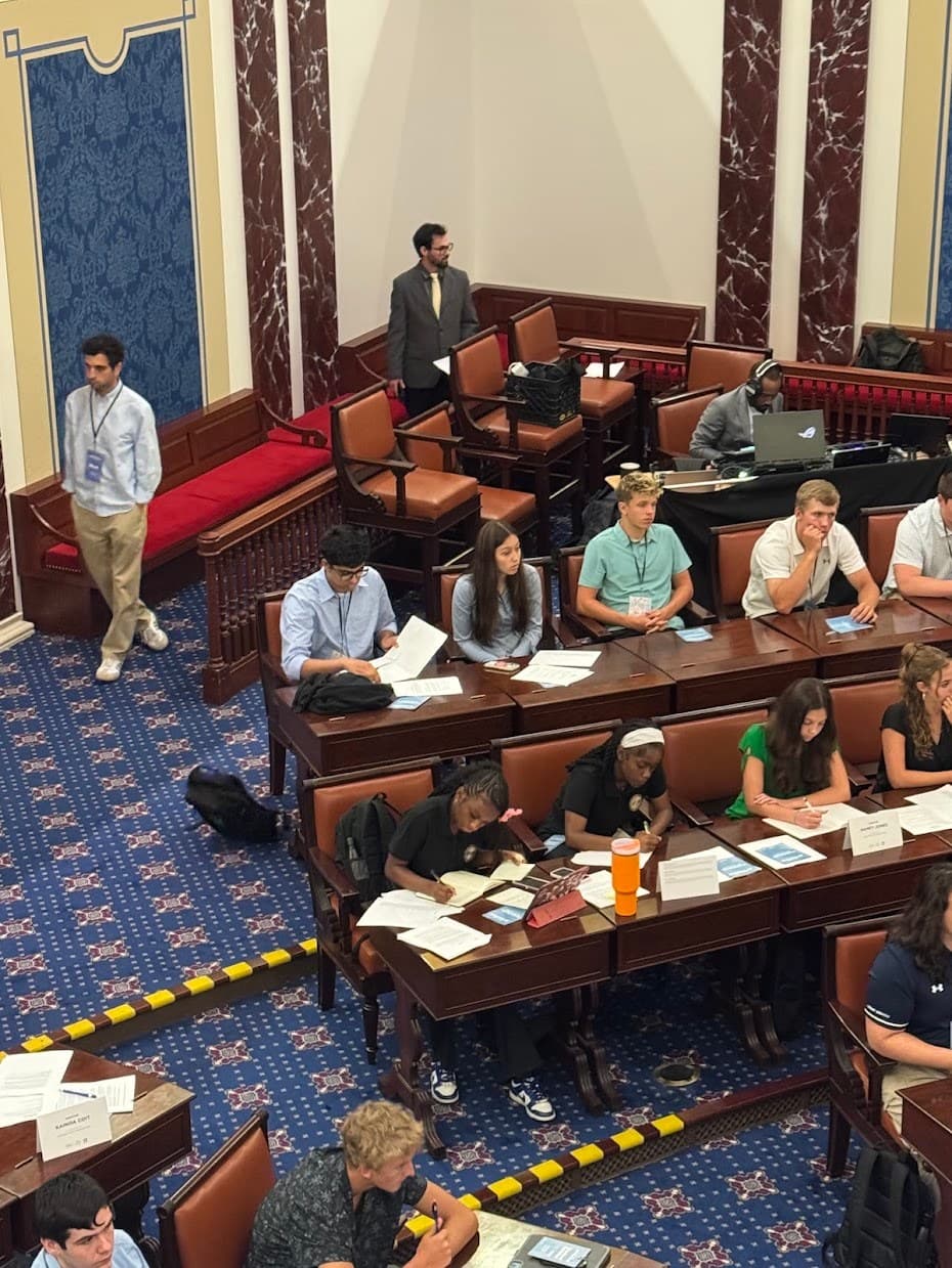 Students seated at desks in a Senate-style chamber, taking notes during a policy session.